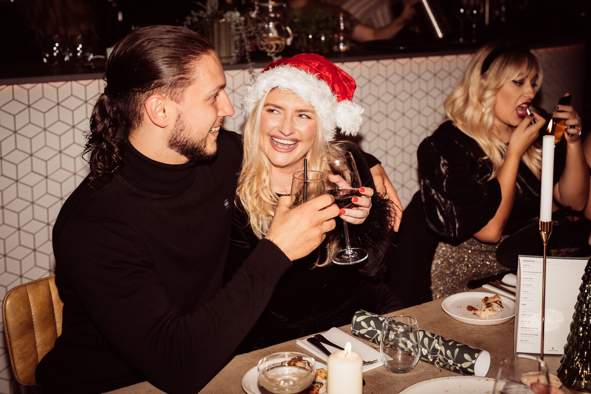 A smiling couple, with the woman wearing a Santa hat, clink wine glasses during a Christmas party meal at Pizza on the Park, a popular Christmas party venue in Bristol. In the background, another guest is enjoying a bite of pizza. The festive table setting includes candles, holiday crackers, and wine, making it an ideal spot for a cozy office Christmas party or work gathering in Clifton.