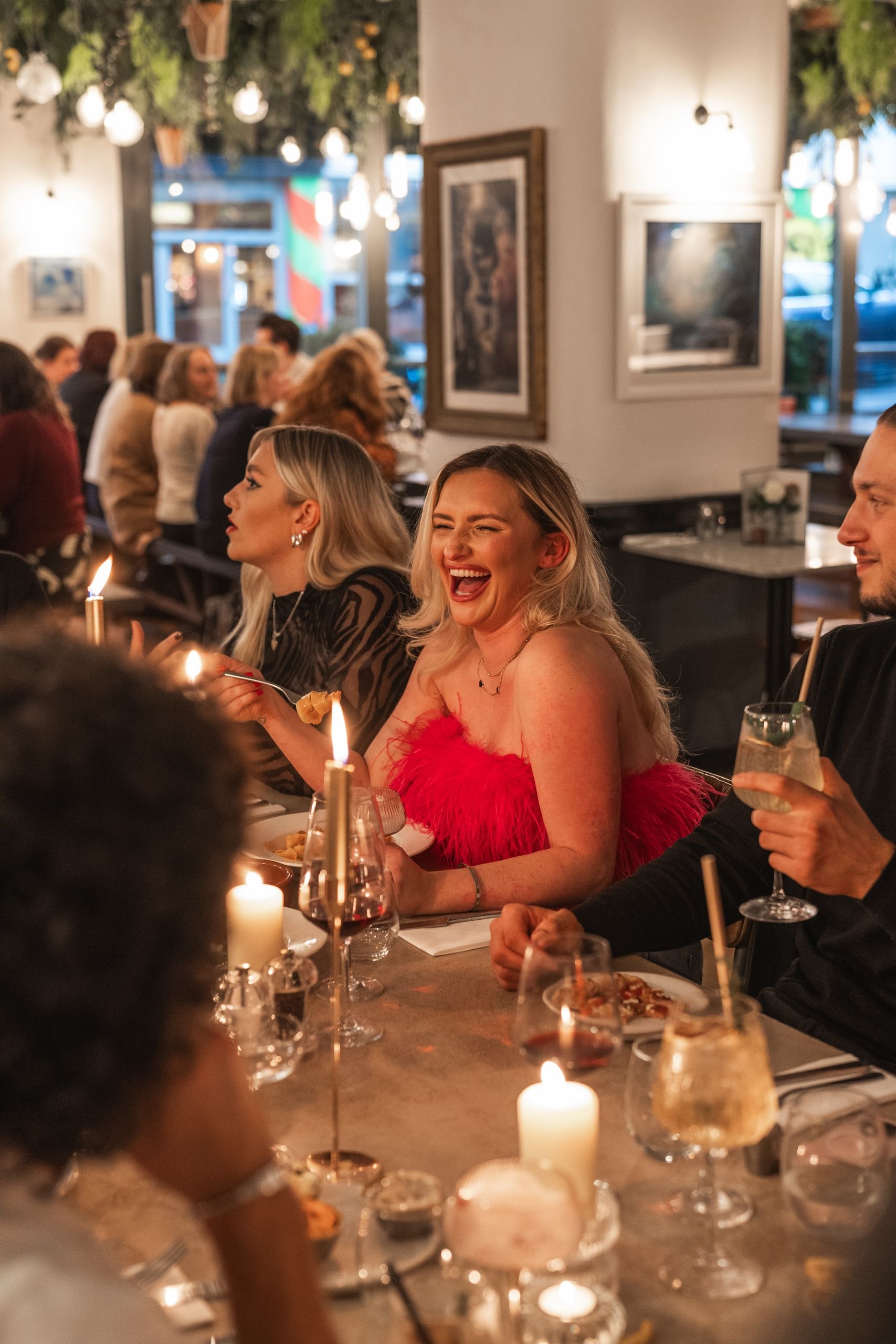 A woman in a red feathered dress laughs with friends over pizza and cocktails at a Christmas party in Pizza on the Park, Clifton. The candlelit setting and festive mood make this the perfect venue for Christmas parties in Bristol, whether for work colleagues or social gatherings.