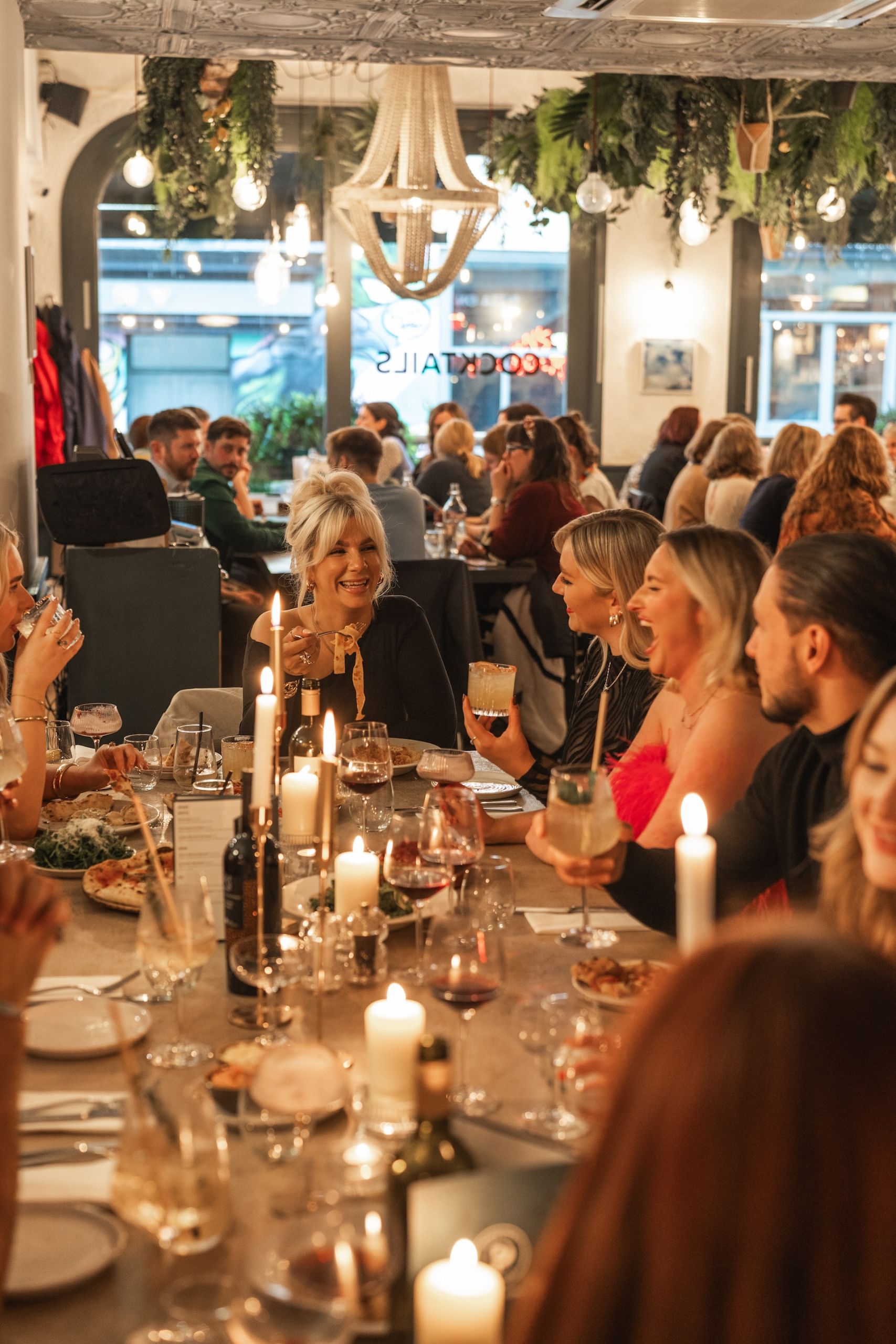 A large group of friends and colleagues, dressed for the festive season, sit at a long table filled with wine, pizza, and cocktails. Candles and holiday crackers complete the Christmas scene at Pizza on the Park, a sought-after Christmas party venue in Bristol. The setting is ideal for office Christmas parties or large group Christmas meals.